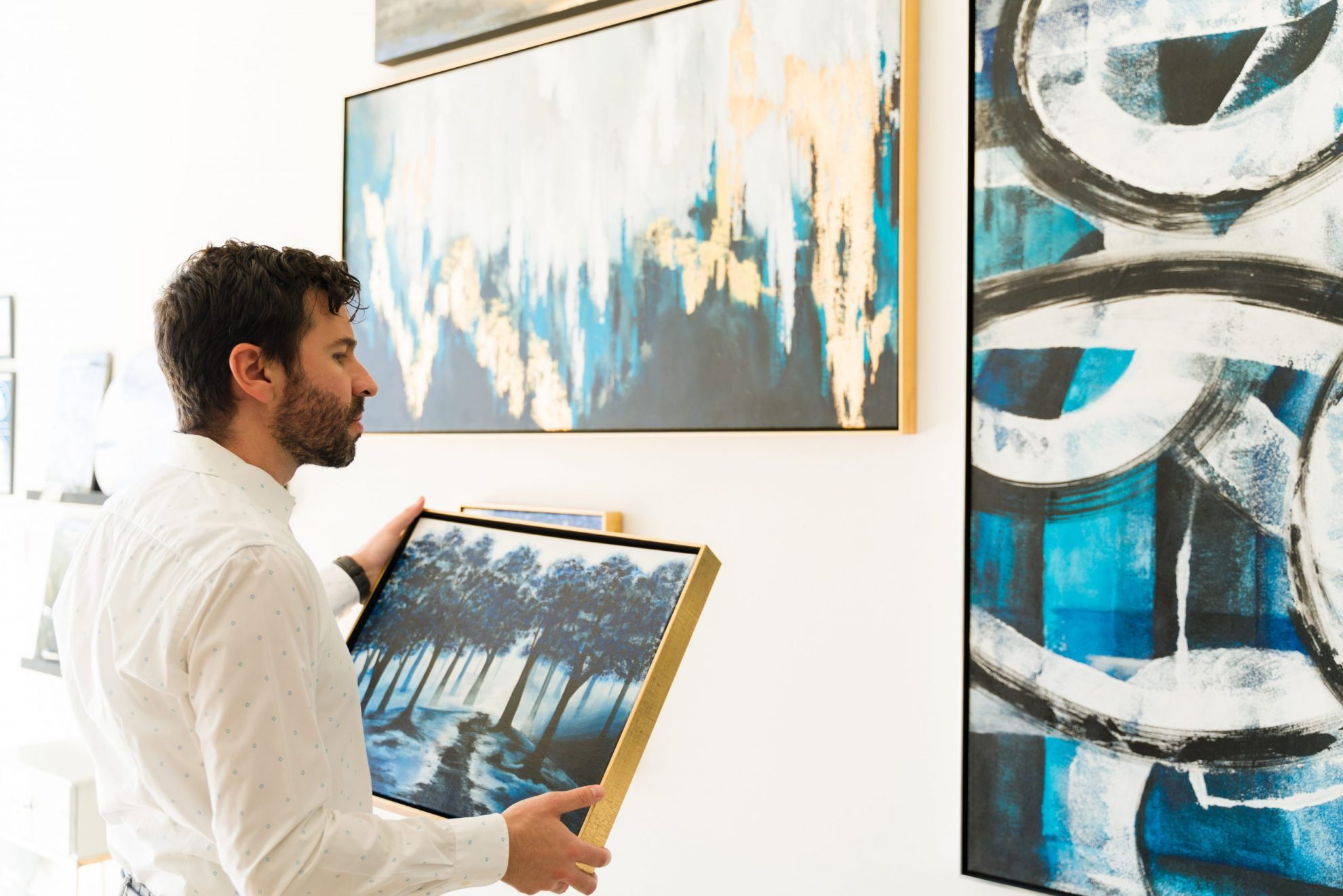 Photo of man holding a painting and looking at artworks illustrates blog; "A Guide to Pawning, Buying, and Selling Paintings and Other Artworks in a Pawn shop"