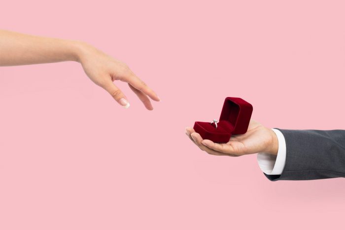 Photo with closeup of hands where a man presents engagement ring to fiancée.