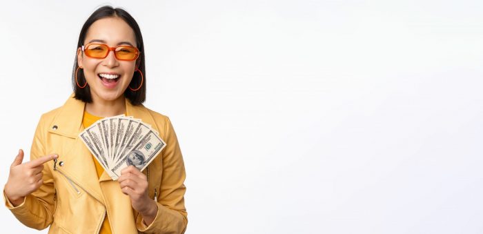 Microcredit and money concept. Stylish asian young woman in sunglasses, laughing happy, holding dollars cash, standing over white background Woman with sunglasses and yellow jacket holding dollar bills illustrates blog "Is Pawning a Loan?"